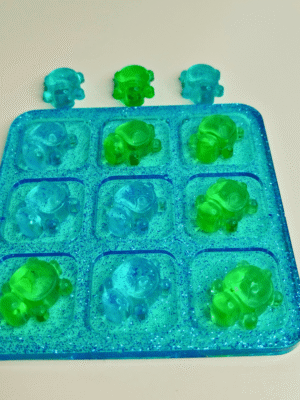 jeu-morpion-resine-tortue Un petit plateau de jeu de morpion de voyage en résine transparente, posé sur une table en bois, illuminé par des inclusions de paillettes bleues scintillantes.