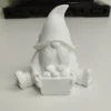 gnome-assis-resine-acrylique-blanc Notre gnome assis, vu de face, montrant son grand bonnet texturé et sa longue barbe, tenant tendrement son petit panier rempli de trois pommes.