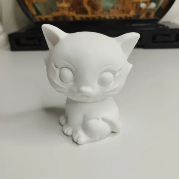 Une statuette chat kawaii à peindre en résine blanc crème, vue de face, posée sur un bureau en bois baigné de lumière naturelle