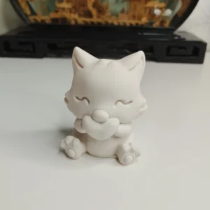 Petite figurine d'un chat blanc assis tenant tendrement un cœur entre ses pattes avant, posée sur une table en bois clair.