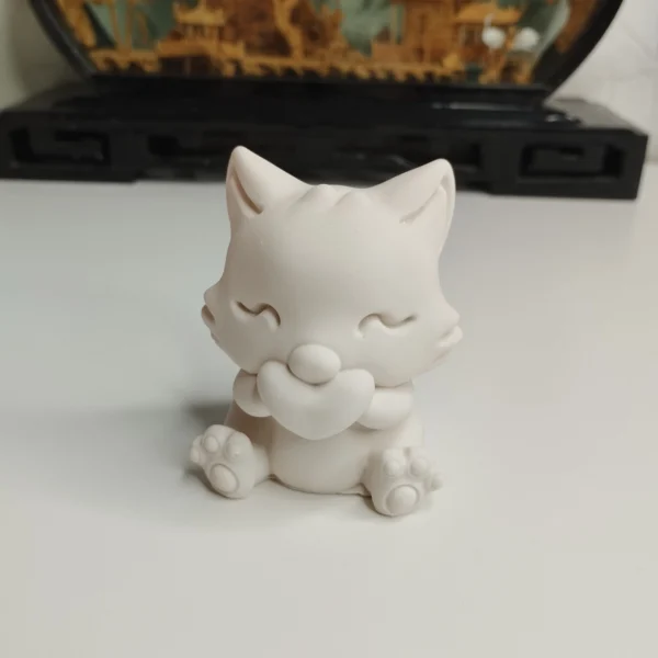 statuette-chat-assis-coeur-face Petite figurine d'un chat blanc assis tenant tendrement un cœur entre ses pattes avant, posée sur une table en bois clair.