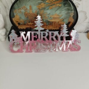 Panneau "Merry Christmas" en résine époxy rose poudré et argenté, posé sur une cheminée avec des bougies.