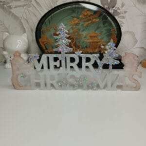 Panneau "Merry Christmas" en résine époxy argentée posé sur une étagère, entouré de bougies LED.