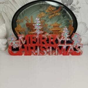 Décoration "Merry Christmas" en résine époxy rouge vif avec paillettes argentées posée sur une étagère en bois