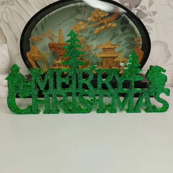 decoration-merry-christmas-resine-face Vue de face d'une décoration de Noël verte en résine, affichant l'inscription Merry Christmas avec des flocons et des sapins scintillants.
