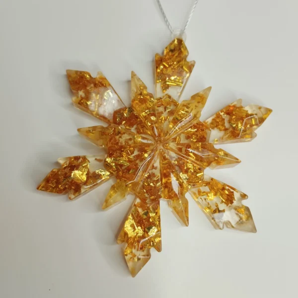 Un grand flocon à huit branches transparent rempli de paillettes dorées, suspendu délicatement à une branche de sapin de Noël.