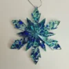 ornement-flocon-neige-resine-face Vue de face d'une décoration sapin flocon de neige en résine, montrant les reflets scintillants bleus et turquoise sur un fond blanc lumineux.