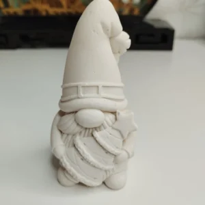 Un adorable gnome de Noël à peindre en résine blanche, photographié de face. Il est assis, porte un grand chapeau pointu qui cache ses yeux, et tient fermement un petit sapin de Noël contre son ventre potelé.