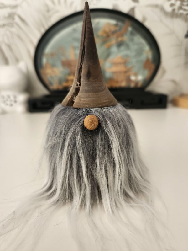 gnome-decoratif-bois-tourne Un adorable gnome décoratif en bois tourné, posé sur une surface plane. On distingue parfaitement son grand chapeau sombre, son petit nez rond en bois clair et sa longue barbe grise qui tombe souplement.