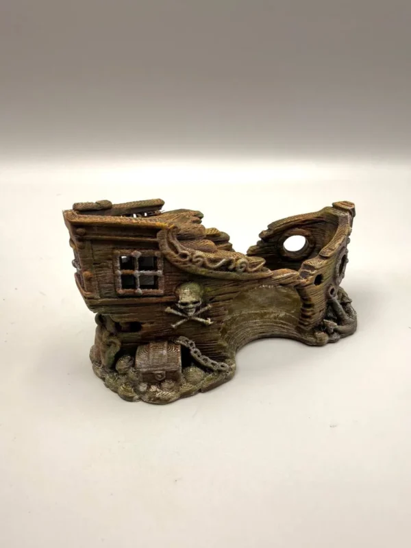 Vue détaillée du socle d'un décor fond marin pour terrarium posé sur un sol texturé, dévoilant un petit coffre aux trésors fermé et entouré de chaînes sculptées à la base de la coque.